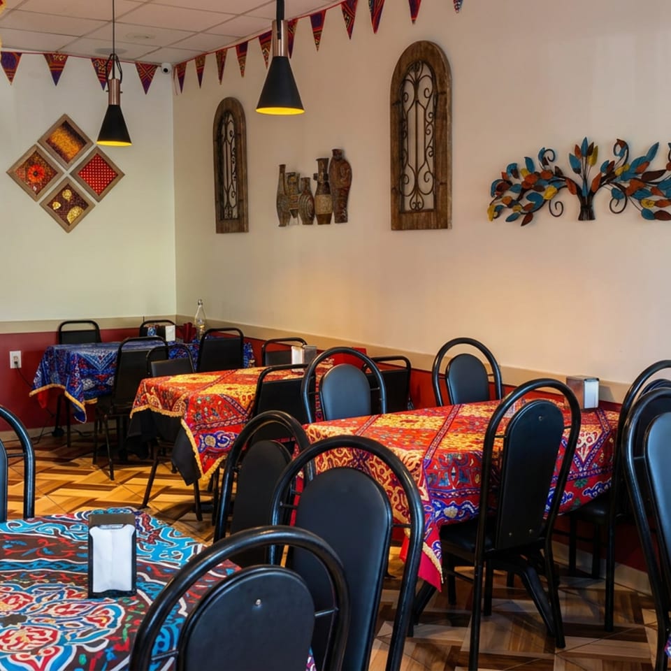 Colorful patterned tables and black chairs fill a warmly lit restaurant decorated with ornate wall art and festive bunting.