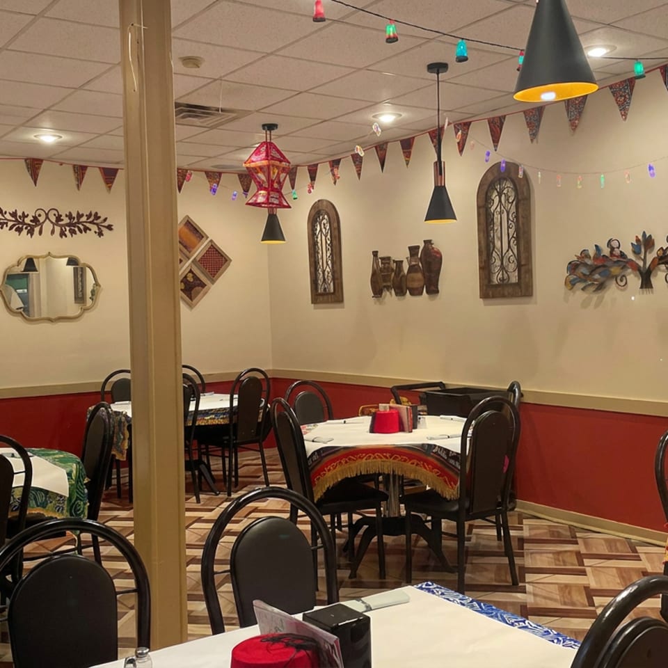 Colorfully decorated restaurant dining room with patterned tablecloths, black chairs, pendant lights, and ornate wall art.